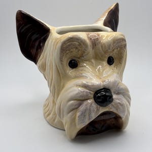 May include: A ceramic mug shaped like the head of a terrier dog. The mug features a cream-colored coat with brown accents on the ears, around the eyes, and on the muzzle. It has a glossy finish, black eyes, and a black nose, with an open top.