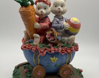 May include: A decorative Easter figurine featuring two white bunnies riding in a blue cart. One bunny holds a large orange carrot, the other an Easter egg. The cart is decorated with flowers and sits on a green base with a miniature railroad track.