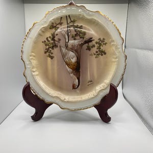 May include: A decorative porcelain plate with a hand-painted image of a bird hanging upside down from a tree branch. The plate has a scalloped edge with gold trim and is displayed on a dark wooden stand.