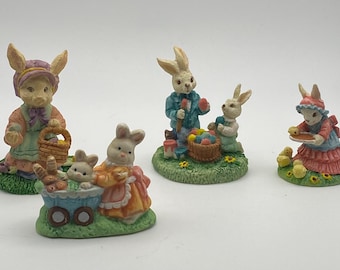 May include: Four Easter bunny figurines. One bunny wears a bonnet and apron, holding a basket. Another bunny figure is pushing a blue cart with two bunnies inside. A third bunny figure is painting eggs with a helper bunny. The last bunny figure is holding a plate with chicks.