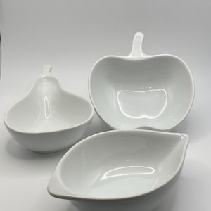 May include: Three white ceramic serving bowls in different shapes. One is pear-shaped, another is apple-shaped with a stem, and the third is leaf-shaped. All bowls have a glossy finish and are set against a white background.
