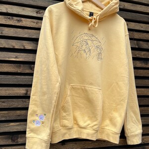 May include: A pale yellow hooded sweatshirt featuring a dark blue line art embroidery on the chest of two figures under an umbrella in the rain. The right sleeve includes a small embroidered detail with two speech bubbles containing the text Hi.