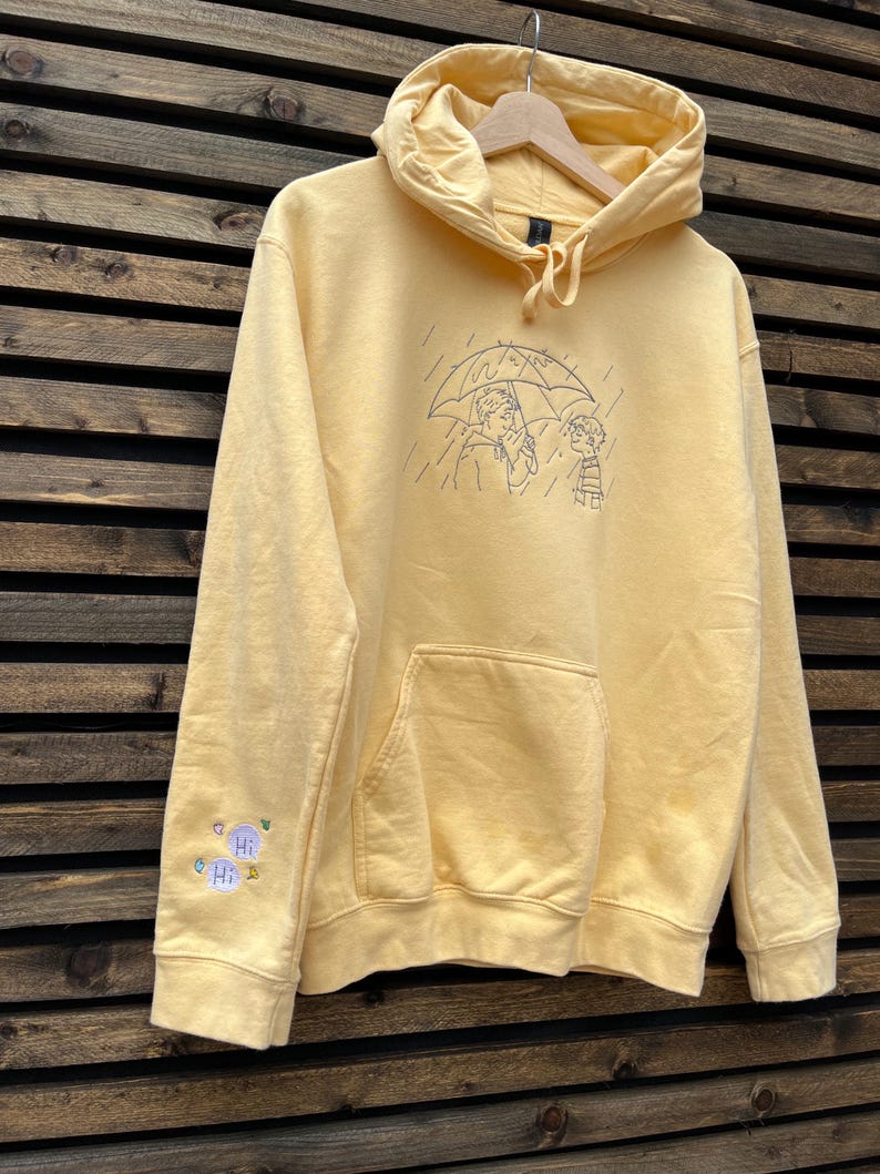 May include: A pale yellow hooded sweatshirt featuring a dark blue line art embroidery on the chest of two figures under an umbrella in the rain. The right sleeve includes a small embroidered detail with two speech bubbles containing the text Hi.