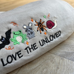 Op de afbeelding: Een lichtgrijs t-shirt met geborduurde ontwerpen van een zwarte vleermuis, een groene kikker, een grijze muis, een oranje en zwarte spin, en een slak met een roze huisje. De tekst "LOVE THE UNLOVED" is in het zwart onder de dieren geborduurd.
