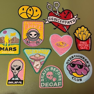 Può includere: Una collezione di toppe ricamate con vari disegni. Include una faccia gialla e nera triste/felice, un cuore rosso con forbici, un cuore rosa con un occhio, un cartone di patatine fritte e toppe con temi alieni, spaziali e teschio.