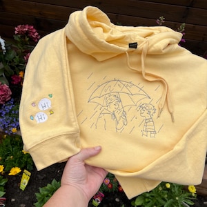 May include: A pale yellow hooded sweatshirt featuring a dark blue embroidered line art design of two figures standing under an umbrella in the rain. The sleeve includes two small speech bubbles with the text Hi and small colorful leaf icons.