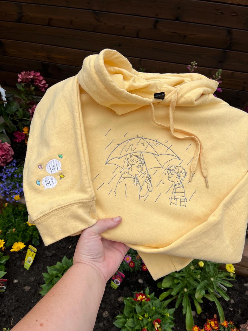 May include: A pale yellow hooded sweatshirt featuring a dark blue embroidered line art design of two figures standing under an umbrella in the rain. The sleeve includes two small speech bubbles with the text Hi and small colorful leaf icons.