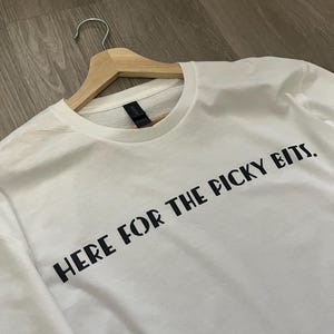 Op de afbeelding: Een wit t-shirt met de zwarte tekst "HERE FOR THE PICKY BITS.". Het t-shirt hangt aan een houten kleerhanger. De achtergrond is een lichtbruin houten oppervlak. Het shirt heeft een ronde hals en korte mouwen.