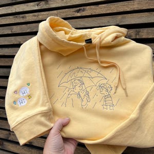 May include: A pale yellow hooded sweatshirt featuring dark blue embroidered line art on the front showing two people under an umbrella in the rain. The right sleeve features two small white speech bubbles with the text Hi and colorful leaf icons.
