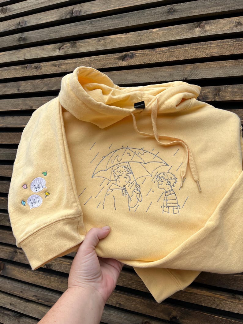May include: A pale yellow hooded sweatshirt featuring dark blue embroidered line art on the front showing two people under an umbrella in the rain. The right sleeve features two small white speech bubbles with the text Hi and colorful leaf icons.