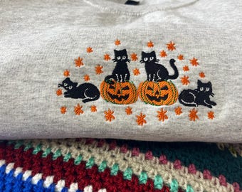 May include: A light gray sweatshirt with an embroidered Halloween design. The design features four black cats, two sitting atop carved orange pumpkins, and orange star accents.