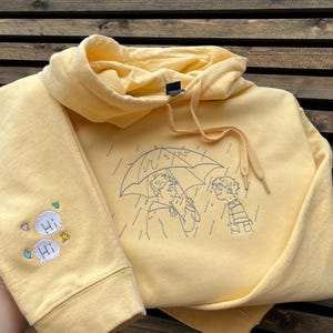 May include: A pale yellow hooded sweatshirt featuring a dark blue embroidered line art illustration of two people standing under an umbrella in the rain. The sleeve displays two embroidered speech bubbles with the text Hi and small colorful accents.