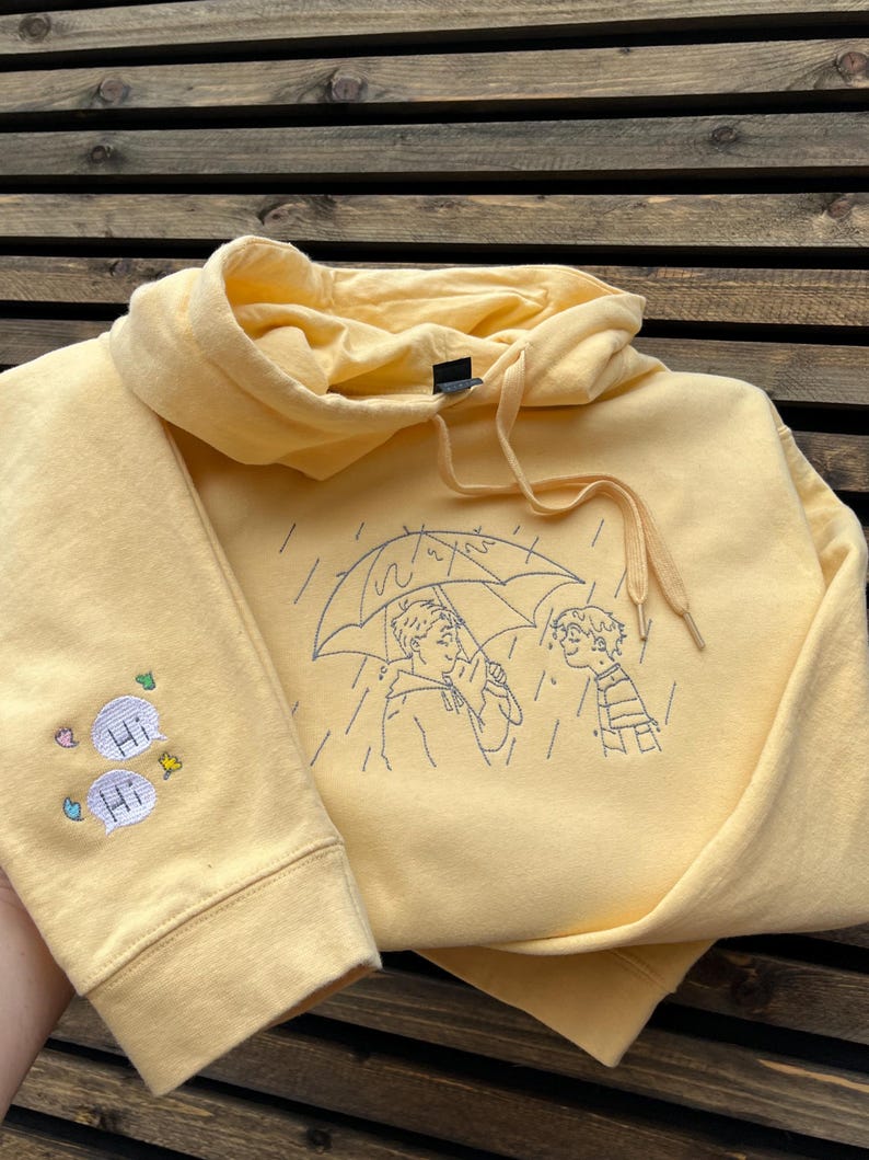 May include: A pale yellow hooded sweatshirt featuring a dark blue embroidered line art illustration of two people standing under an umbrella in the rain. The sleeve displays two embroidered speech bubbles with the text Hi and small colorful accents.