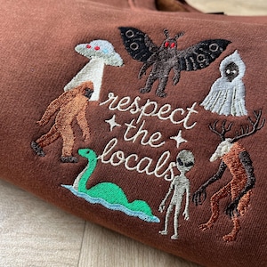 May include: A brown sweatshirt featuring embroidered cryptid figures including a mothman, bigfoot, a ghost, a green sea creature, an alien, and a deer-like creature. The text "respect the locals" is embroidered in white in the centre.