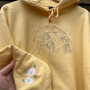 May include: A pale yellow hooded sweatshirt featuring dark thread embroidery on the chest depicting two figures under an umbrella in the rain. The sleeve includes embroidered speech bubbles with the text "Hi" and small colorful leaf and flower accents.