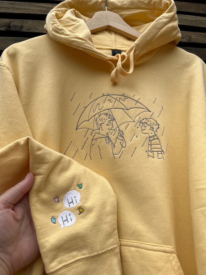 May include: A pale yellow hooded sweatshirt featuring dark thread embroidery on the chest depicting two figures under an umbrella in the rain. The sleeve includes embroidered speech bubbles with the text "Hi" and small colorful leaf and flower accents.