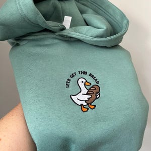 Könnte beinhalten: Ein salbeigrüner Kapuzenpullover mit einer Stickerei, die eine weiße Ente mit orangefarbenem Schnabel und Füßen zeigt, die einen braunen Laib Brot hält. Der Text "LETS GET THIS BREAD" ist in einem Bogen über der Ente in schwarzem Garn gestickt.