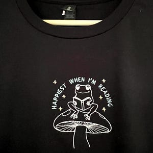 Può includere: T-shirt nera con un disegno bianco di una rana che legge un libro su un fungo. Il testo "Happiest When I'm Reading" circonda l'immagine. La t-shirt è appesa a una gruccia di legno.