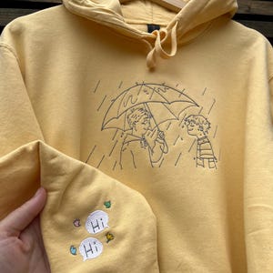 May include: A yellow hooded sweatshirt featuring black line embroidery of two people under an umbrella in the rain. The sleeve is embroidered with two white speech bubbles containing the text "Hi" and small colorful leaf and flower accents.