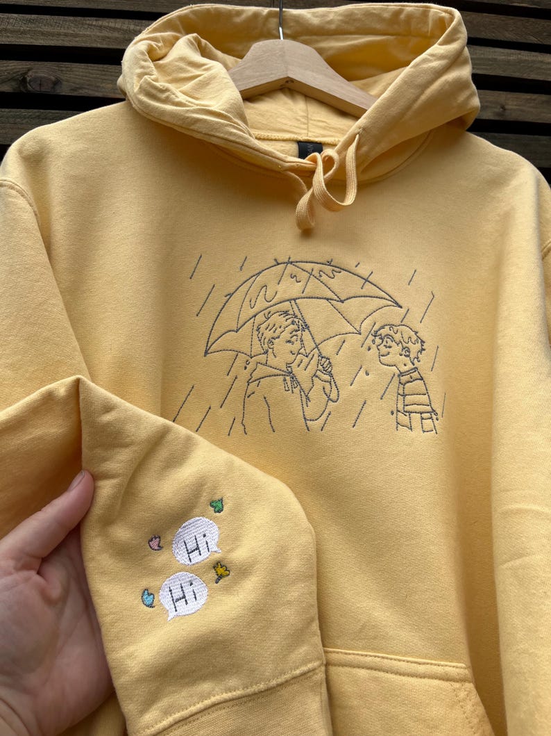 May include: A yellow hooded sweatshirt featuring black line embroidery of two people under an umbrella in the rain. The sleeve is embroidered with two white speech bubbles containing the text "Hi" and small colorful leaf and flower accents.