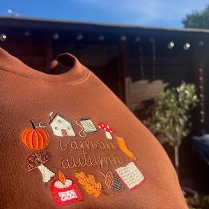 May include: A brown crewneck sweatshirt featuring an embroidered design with the text "I am an autumn" surrounded by icons of a pumpkin, a house, a spool of thread, mushrooms, a candle, an oak leaf, an acorn, and an open book.