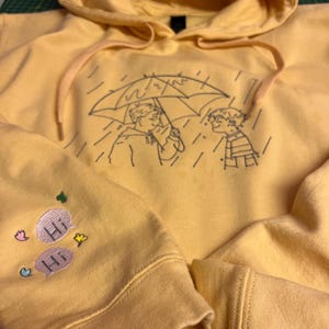 May include: A bright yellow hooded sweatshirt featuring a black embroidered line art illustration of two figures under an umbrella in the rain. The sleeve displays two embroidered speech bubbles with the text Hi, accented by small colorful hearts and stars.