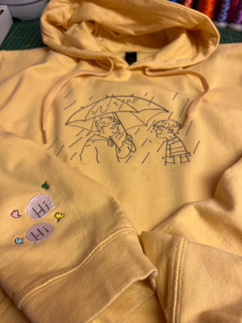 May include: A bright yellow hooded sweatshirt featuring a black embroidered line art illustration of two figures under an umbrella in the rain. The sleeve displays two embroidered speech bubbles with the text Hi, accented by small colorful hearts and stars.
