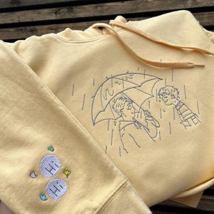 May include: A pale yellow hooded sweatshirt featuring dark blue line art embroidery of two people under an umbrella in the rain. The sleeve displays two light purple speech bubbles with the text "HI" embroidered inside, accented by small colorful shapes.