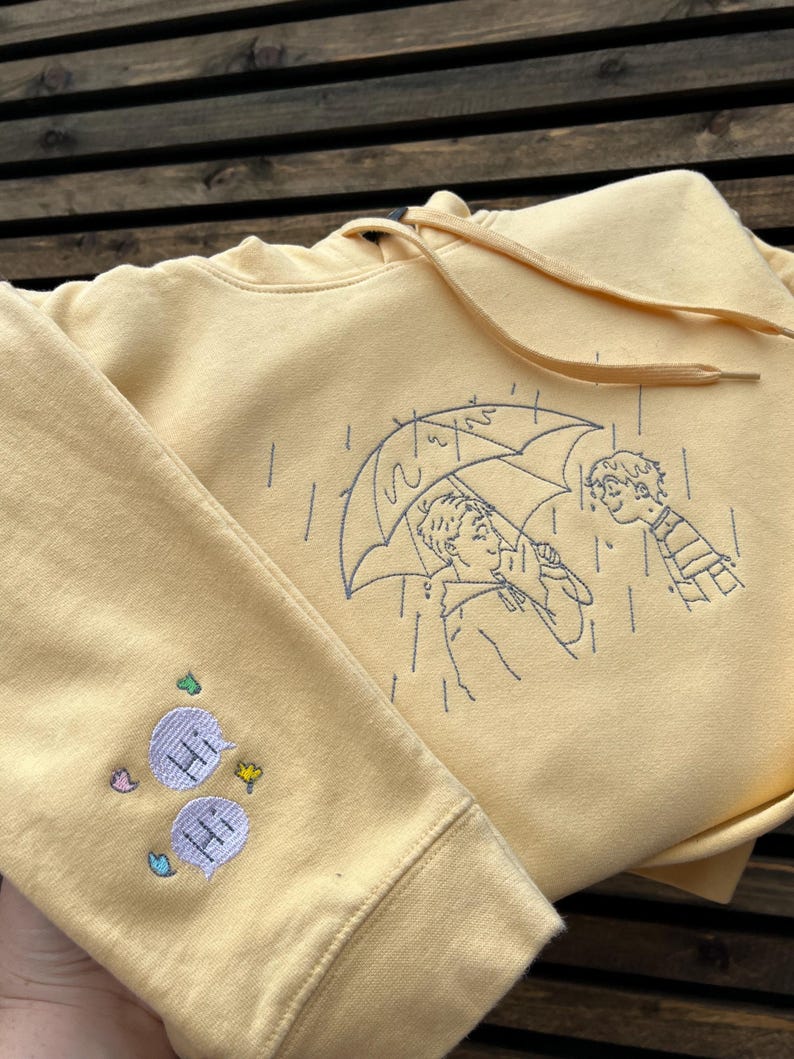 May include: A pale yellow hooded sweatshirt featuring dark blue line art embroidery of two people under an umbrella in the rain. The sleeve displays two light purple speech bubbles with the text "HI" embroidered inside, accented by small colorful shapes.