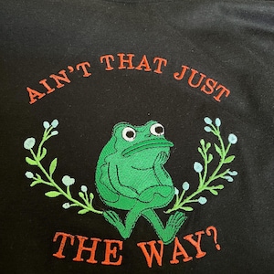 May include: Black sweatshirt with an embroidered design. It features a green frog with a thoughtful expression, surrounded by a floral wreath. The text "AIN'T THAT JUST THE WAY?" is embroidered in orange.