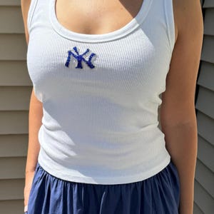 Op de afbeelding: Witte geribbelde tanktop met een blauw geborduurd "NY" logo. De tanktop heeft een ronde hals en wordt gecombineerd met een marineblauwe rok. De geribbelde textuur geeft een klassieke uitstraling aan de casual outfit.