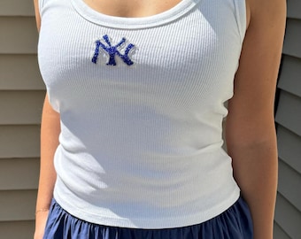 May include: White ribbed tank top with a blue embroidered "NY" logo. The tank top has a scoop neckline and is paired with a navy blue skirt. The ribbed texture adds a classic touch to the casual outfit.