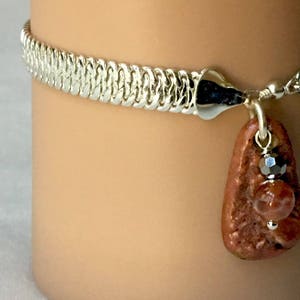 May include: A silver chain bracelet with a brown stone pendant. The pendant has a silver bead and a smaller red stone.