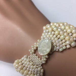 May include: A white beaded bracelet with a large, off-white oval-shaped stone in the center. The bracelet is made of small, iridescent beads and has a delicate, feminine look.