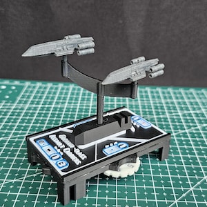 Op de afbeelding: Een miniatuur grijs ruimteschipmodel met twee ruimteschepen bevestigd aan een zwarte standaard. De standaard is gemonteerd op een zwarte basis met witte en blauwe details. De basis heeft de tekst "Ancient Quasar".