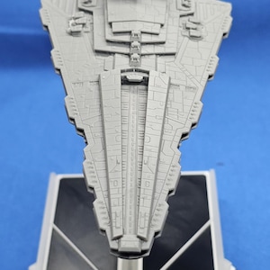 Könnte beinhalten: Ein graues Modell eines Venator-Klasse Sternenzerstörers, ein Raumschiff aus dem Star Wars-Universum, wird auf einem schwarz-weißen Ständer präsentiert. Das Modell weist detaillierte Oberflächenmerkmale auf und ist vor einem blauen Hintergrund platziert. Der Ständer trägt den Text "Venator II-Class Star Destroyer".