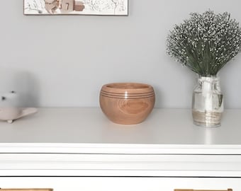 May include: A light wood bowl with dark brown stripes around the rim sits on a white dresser with two drawers. The dresser has gold handles. A bouquet of white flowers in a clear glass vase is on the dresser to the right of the bowl.