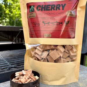 May include: A bag of cherry BBQ smoking chips with the text "CHERRY" and "100% NATURAL". The bag is partially transparent, revealing wood chips. A small, round, metal container filled with wood chips sits in front of the bag.