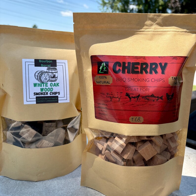 May include: Two kraft paper bags of wood smoker chips. One bag has a label that reads "WHITE OAK WOOD SMOKER CHIPS". The other bag has a red label that reads "CHERRY BBQ SMOKING CHIPS" and "1 LB". The chips are visible through the clear window.