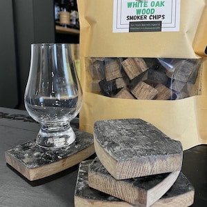 May include: A set of four brown wooden coasters made from white oak barrels. The coasters are stacked on top of each other. A glass of whiskey sits on top of the coasters. A bag of white oak wood smoker chips is in the background.