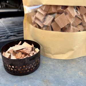 May include: A metal smoker box filled with wood chips next to a bag of wood chunks. The wood chips and chunks are various shades of brown. The smoker box has a perforated design.