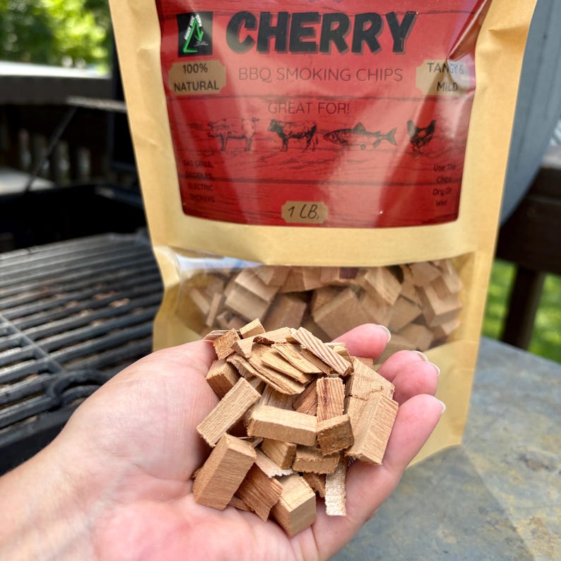 May include: A hand holds a pile of cherry wood BBQ smoking chips. The chips are light brown and rectangular. A bag of chips is in the background, with the text "CHERRY BBQ SMOKING CHIPS" and "1 LB". The bag also says "100% NATURAL".