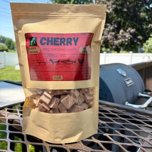 May include: A bag of cherry BBQ smoking chips. The bag is tan with a clear window showing the wood chips. The label is red with white text that reads "CHERRY BBQ SMOKING CHIPS" and "1 LB.". The bag is sitting on a metal grill.