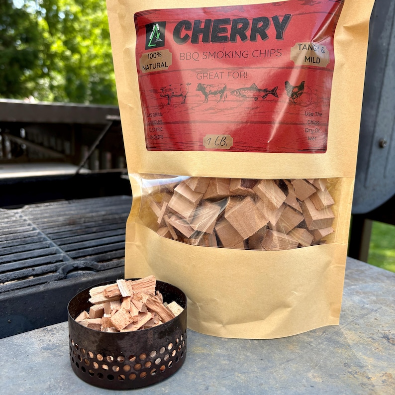 May include: A bag of cherry BBQ smoking chips with the text "CHERRY" and "100% NATURAL". The bag is partially transparent, revealing wood chips. A small, round, metal container filled with wood chips sits in front of the bag.