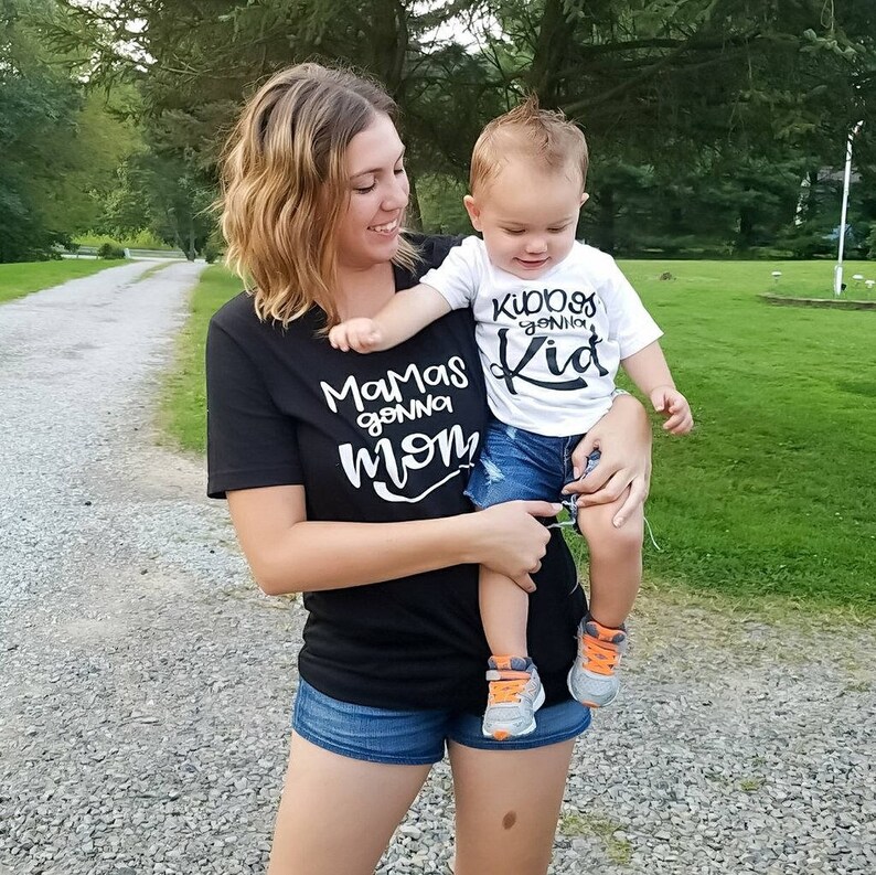 Op de afbeelding: Een vrouw draagt een zwart T-shirt met de tekst "Mamas gonna mom" in witte letters en houdt een jong kind vast dat een wit T-shirt draagt met de tekst "Kiddos gonna Kid" in zwarte letters. De vrouw draagt een blauwe spijkerbroek en het kind draagt een blauwe spijkerbroek.