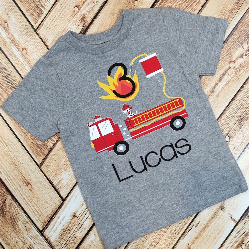 Op de afbeelding: Grijs T-shirt met korte mouwen met een rode brandweerwagen, een dalmati&euml;r en een gele vlam met het nummer 3. De tekst "Lucas" is onder de brandweerwagen gedrukt.