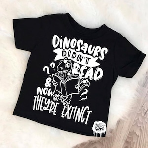 May include: Black t-shirt with a white graphic of a dinosaur reading a book. The text reads "Dinosaurs didn't read & now they're extinct".