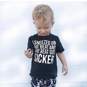 May include: A black t-shirt with white text that reads "I sneezed on the beat and the beat got Sicker". A young child is wearing the shirt and looking down.