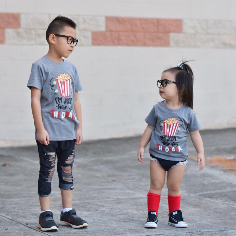 May include: Two children wearing grey t-shirts with a popcorn bucket graphic and the text "I'm just here for the snacks". The boy is wearing black ripped jeans and the girl is wearing a white lace diaper cover and red knee-high socks. Both children are wearing black framed glasses.