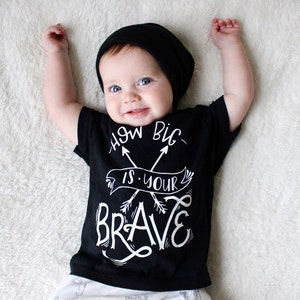 Op de afbeelding: Een zwart T-shirt met witte tekst die "How Big Is Your Brave" zegt met twee pijlen die in het midden kruisen. Een baby draagt het shirt en een zwarte beanie.